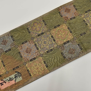 May include: A decorative textile with a repeating pattern of squares. The squares alternate between a wood grain design and a floral pattern. The colors include shades of brown, green, gold, and gray. There is Japanese text on a small rectangular patch.