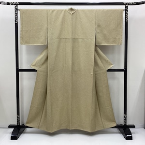 May include: A beige kimono displayed on a black metal rack. The kimono has a subtle pattern and is draped to show the sleeves and the front panels. The rack has decorative elements on the top and is set against a white background.