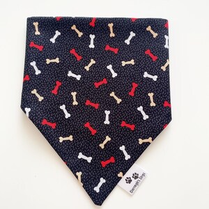 May include: Black bandana with a white and tan bone pattern and red bone accents. The bandana has a small tag that says "Darragh's Dogs" with a paw print logo.
