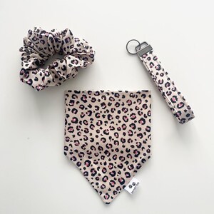 May include: A set of three dog accessories in a leopard print pattern. The set includes a bandana, a scrunchie, and a key chain. The bandana has a pink and black leopard print on a beige background. The scrunchie is made of a soft, stretchy fabric. The key chain is made of a durable fabric and has a metal ring.