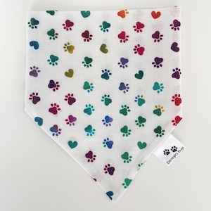 May include: White dog bandana featuring a pattern of colourful paw prints and hearts. The paw prints and hearts are in shades of red, purple, blue, green, and yellow. A small tag in the corner reads "Darragh's Dogs".