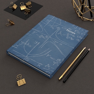 May include: A blue notebook with a white mathematical equation pattern. The notebook has the word "Notes" written on the cover. Two pencils, one gold and one black, are lying on a brown leather surface next to the notebook.