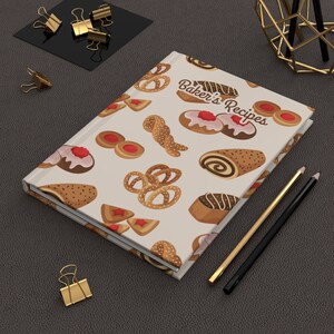 May include: A cream-coloured notebook with a pattern of various pastries and the text "Baker's Recipes". The notebook is lying on a brown leather surface with two pencils and gold paper clips.