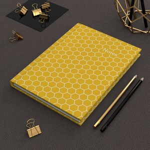 May include: A yellow notebook with a white honeycomb pattern and the word "Notes" printed on the cover. The notebook is lying on a dark brown surface with gold paper clips and pencils.
