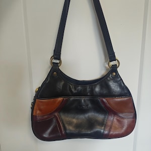 Puede incluir: Un bolso de hombro con un diseño de bloques de color en negro, marrón y burdeos intenso. El bolso tiene una parte superior curva, dos correas largas para el hombro unidas con anillos de metal dorado y la marca La Diva en el panel frontal.