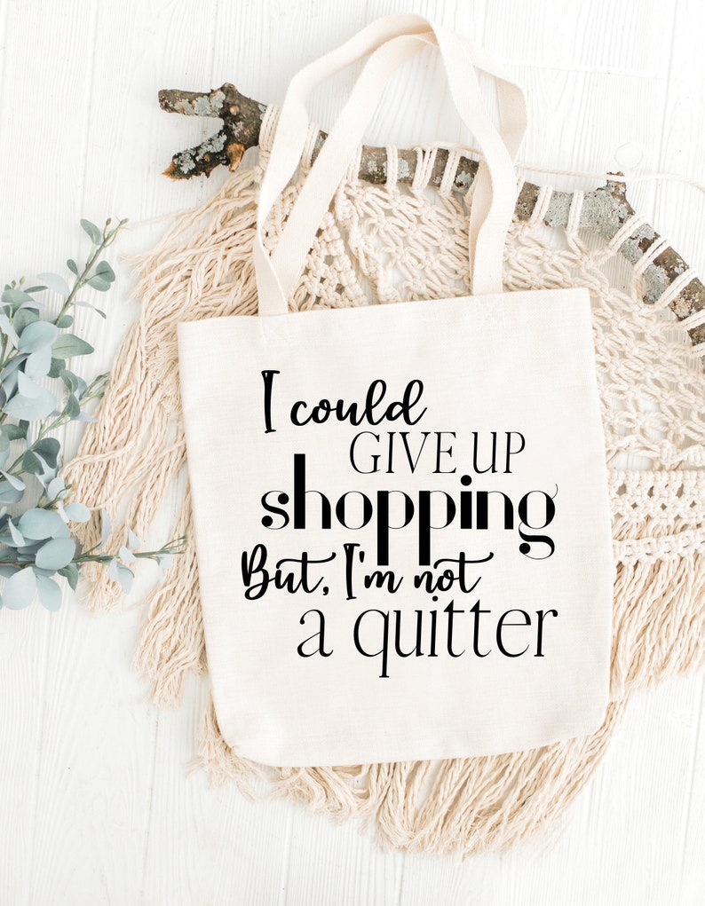 Op de afbeelding: Een witte canvas tote bag met zwarte tekst die "Ik zou kunnen stoppen met winkelen, maar ik ben geen opgever" zegt.
