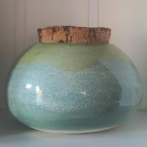 May include: A handmade, round ceramic jar with a cork stopper. The jar is glazed in shades of blue and green, with a speckled texture. The cork stopper is a natural brown color.