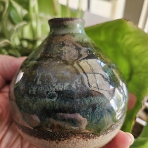 May include: A small, round ceramic vase with a narrow neck and a glossy, multi-colored glaze featuring deep shades of green, blue, and brown. The base of the vase is made of unglazed, textured beige clay, creating a contrast with the smooth, reflective upper portion.