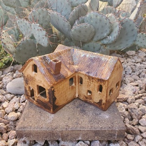 May include: A handcrafted ceramic house sculpture with a brown, textured exterior and a chimney. The roof has a glazed finish. The house sits on a square stone base, with a cactus in the background.