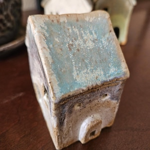 May include: A small ceramic house figurine featuring a textured, light blue glazed roof and a speckled off-white body. The structure includes a small arched doorway and two circular windows on the front facade, with a weathered, rustic finish.
