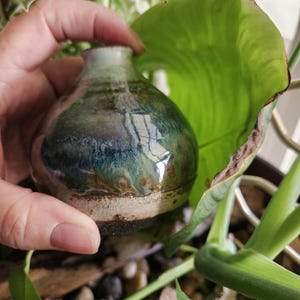 May include: A small, round ceramic vase with a narrow neck, featuring a glossy glaze in shades of deep forest green, teal, and blue, transitioning to a textured, matte beige and dark brown base with visible clay grain and earthy tones.