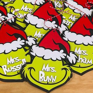 Puede incluir: Adornos verdes y blancos con tem&aacute;tica de Grinch con gorros rojos de Pap&aacute; Noel. Cada adorno tiene un nombre diferente escrito en &eacute;l, incluyendo "Mrs. Russell", "Mrs. Bunn" y "Mrs. Graham".