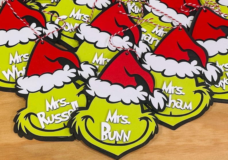 Puede incluir: Adornos verdes y blancos con tem&aacute;tica de Grinch con gorros rojos de Pap&aacute; Noel. Cada adorno tiene un nombre diferente escrito en &eacute;l, incluyendo "Mrs. Russell", "Mrs. Bunn" y "Mrs. Graham".