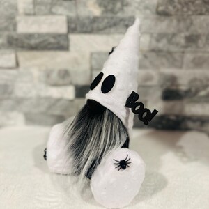 May include: A Halloween gnome dressed as a ghost. The white gnome has a pointed hat with black eyes, a long gray beard, and a small spider on its body. A black sign that says "Boo!" is attached to the gnome.