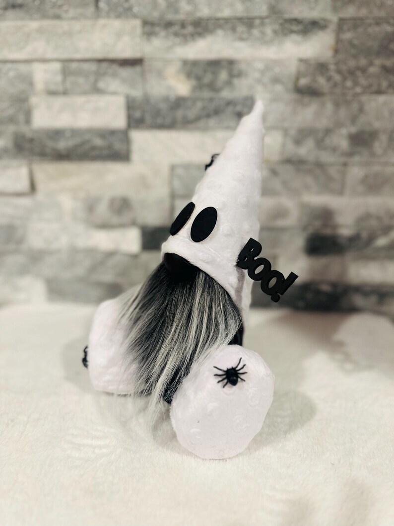 May include: A Halloween gnome dressed as a ghost. The white gnome has a pointed hat with black eyes, a long gray beard, and a small spider on its body. A black sign that says "Boo!" is attached to the gnome.
