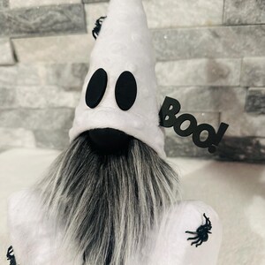 May include: A Halloween-themed gnome with a white ghost costume. The gnome has a long gray and white beard, black eyes, and a black "BOO!" sign. Black spiders are attached to the costume.