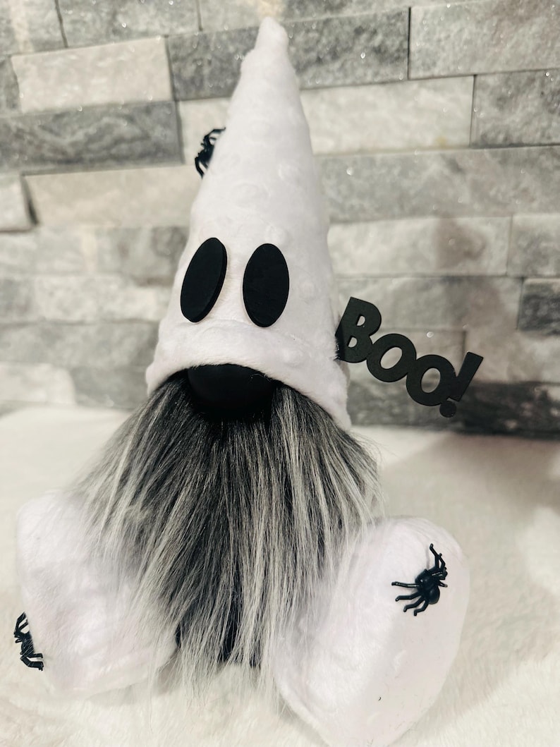 May include: A Halloween-themed gnome with a white ghost costume. The gnome has a long gray and white beard, black eyes, and a black "BOO!" sign. Black spiders are attached to the costume.