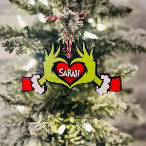 Puede incluir: Adorno de mano verde del Grinch con un coraz&oacute;n rojo que dice "SARAH" en su interior. El adorno est&aacute; colgado de un &aacute;rbol de Navidad verde.