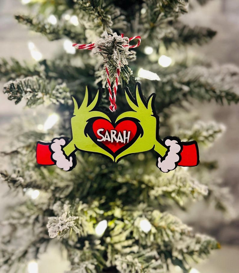 Puede incluir: Adorno de mano verde del Grinch con un coraz&oacute;n rojo que dice "SARAH" en su interior. El adorno est&aacute; colgado de un &aacute;rbol de Navidad verde.