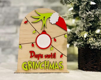 May include: Wooden countdown sign with a green Grinch hand holding a red ornament. The sign says "Days until GRINCHMAS" with green and red lettering.
