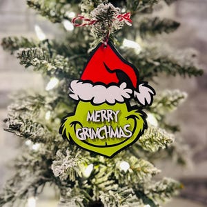 Puede incluir: Adorno navide&ntilde;o verde Grinch con un gorro de Pap&aacute; Noel rojo y las palabras "Merry Grinchmas" en &eacute;l. El adorno est&aacute; colgado de un peque&ntilde;o &aacute;rbol de Navidad artificial.
