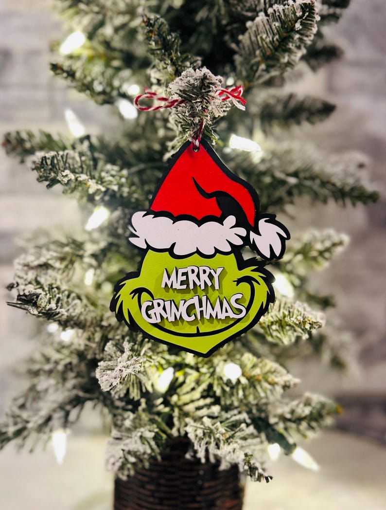 Puede incluir: Adorno navide&ntilde;o verde Grinch con un gorro de Pap&aacute; Noel rojo y las palabras "Merry Grinchmas" en &eacute;l. El adorno est&aacute; colgado de un peque&ntilde;o &aacute;rbol de Navidad artificial.