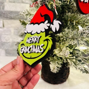 Puede incluir: Adorno navide&ntilde;o verde Grinch con un gorro de Pap&aacute; Noel rojo y las palabras "Merry Grinchmas" en &eacute;l.