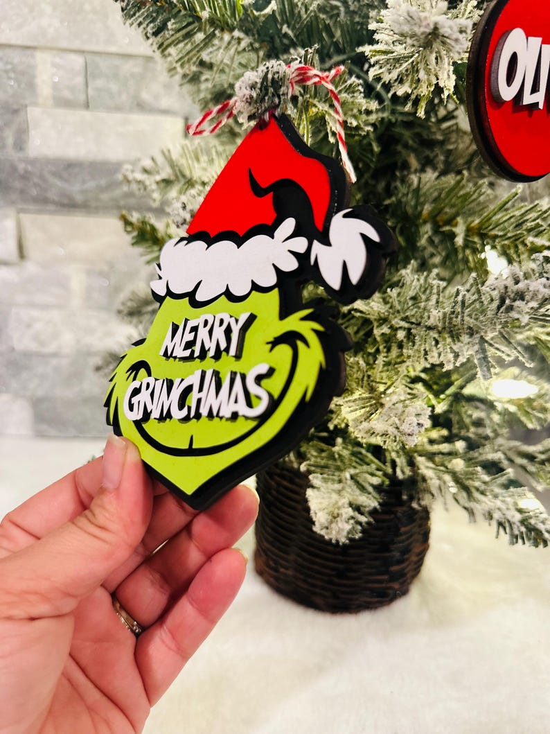 Puede incluir: Adorno navide&ntilde;o verde Grinch con un gorro de Pap&aacute; Noel rojo y las palabras "Merry Grinchmas" en &eacute;l.