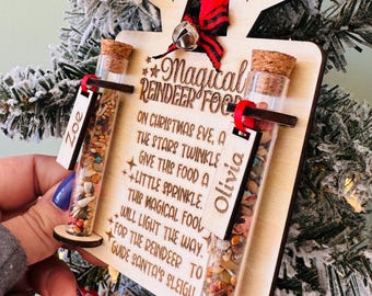 Puede incluir: Adorno de reno de madera con dos tubos de vidrio llenos de chispas de colores. El adorno tiene el texto "Magical Reindeer Food" e instrucciones para usar las chispas en Nochebuena. El adorno está personalizado con los nombres "Zoe" y "Olivia".
