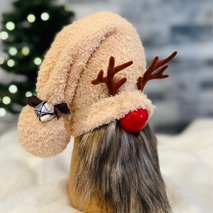 May include: A plush reindeer gnome with a brown and white beard, a red nose, and brown antlers. The gnome is wearing a beige fuzzy hat with a bell.