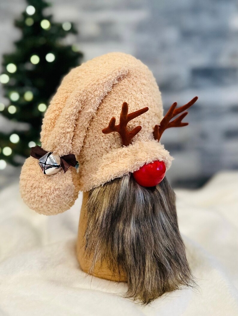 May include: A plush reindeer gnome with a brown and white beard, a red nose, and brown antlers. The gnome is wearing a beige fuzzy hat with a bell.