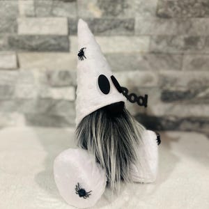 May include: A Halloween gnome with a white hat and body, black eyes, and a long gray beard. The hat and body are decorated with small black spiders. A black sign that says "boo" is attached to the gnome.