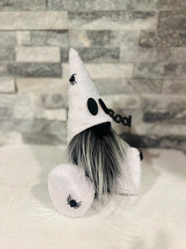 May include: A Halloween gnome with a white hat and body, black eyes, and a long gray beard. The hat and body are decorated with small black spiders. A black sign that says "boo" is attached to the gnome.