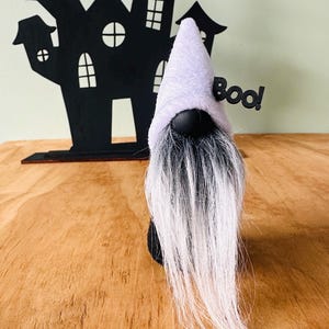 May include: A Halloween gnome with a lavender hat and long white beard stands in front of a black haunted house silhouette. The word "Boo!" is attached to the gnome's hat. The gnome is a Halloween decoration.