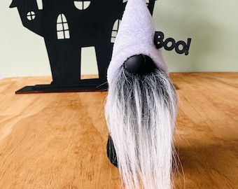 May include: A Halloween gnome with a lavender hat and long white beard stands in front of a black haunted house silhouette. The word "Boo!" is attached to the gnome's hat. The gnome is a Halloween decoration.