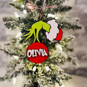 Puede incluir: Mano verde del Grinch sosteniendo un adorno rojo con el nombre Olivia escrito en letras blancas.