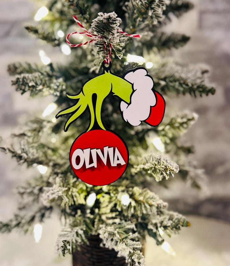 Puede incluir: Mano verde del Grinch sosteniendo un adorno rojo con el nombre Olivia escrito en letras blancas.
