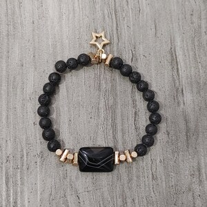 May include: Black lava rock bead bracelet with gold-coloured accents and a rectangular black stone. A gold star charm hangs from the top of the bracelet. The bracelet is on a grey surface.