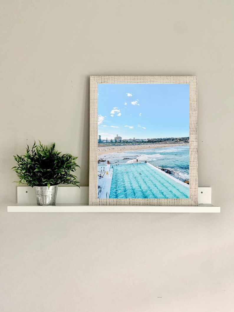 May include: A framed photograph of a swimming pool with a view of the ocean and city skyline. The pool is surrounded by a white tiled deck and there are people swimming in the pool.