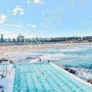 May include: An aerial view of a large outdoor swimming pool with a city skyline in the background. The pool is surrounded by a concrete deck and is filled with clear blue water. The sky is a bright blue with white clouds. There are white outlines of aeroplanes drawn on the sky.