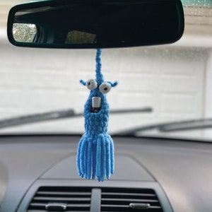 May include: A blue crocheted car air freshener hanging from the rearview mirror of a car. The air freshener has a long, thin body with a round head and two large, white eyes. The body is covered in blue yarn and has a small, white, cylindrical object attached to the head.