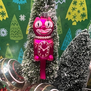 May include: Pink cat-shaped Christmas ornament with a clock face and a pearl necklace. The ornament is hanging on a small artificial Christmas tree.