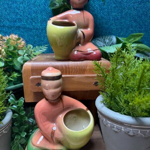 May include: Two ceramic figurines of Asian figures, each holding a yellow-green vase. The figures are dressed in peach-colored robes with black accents and green accents. One figurine is seated on a wooden block, and the other is below it. The background includes greenery.