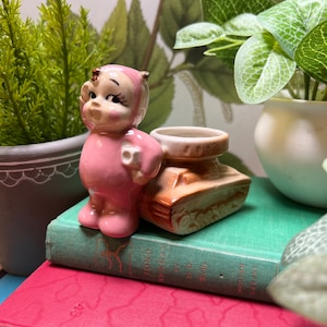 May include: A pink ceramic figurine of a girl wearing a pink dress and holding a cup, standing next to a brown ceramic tank with the word "1000" on it.