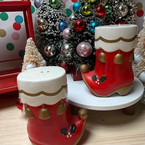 May include: A pair of ceramic Christmas boot salt and pepper shakers. The boots are red with white tops, gold trim, and small gold bells. Green holly and white berries decorate the front of the boots. The shakers are on a white surface with Christmas trees in the background.