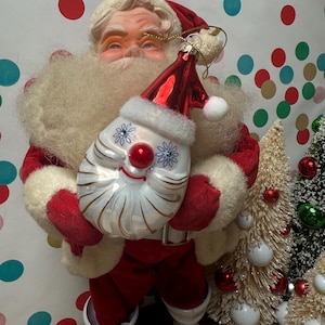 May include: A vintage Santa Claus figure in a red suit and white beard holds a Christmas ornament. The ornament is a smiling face with a red hat. A small Christmas tree with red and white ornaments is in the background.