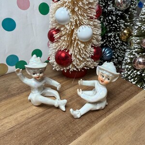 May include: Two vintage ceramic elf figurines with iridescent finish, posed on a wooden surface. The elves wear hats and are in playful positions. Behind them are bottle brush Christmas trees decorated with red and white ornaments.