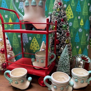 May include: A set of five ceramic Christmas boot mugs, one large pitcher and four smaller mugs, all in white with blue and green accents. The mugs are arranged on a red metal cart with a Christmas tree in the background.