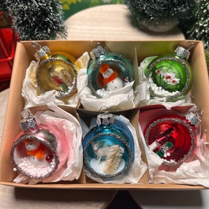 May include: A set of six Christmas ornaments in a cardboard box. Each glass ornament is a different color, including gold, green, pink, blue, and red. The ornaments feature miniature winter scenes with snowmen, deer, and floral designs.