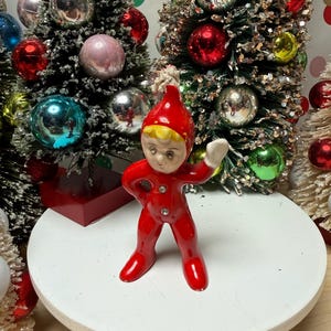 May include: A vintage Christmas figurine of a small elf in a red jumpsuit and hat with a white pom-pom. The elf is standing on a white platform, with Christmas trees and ornaments in the background.
